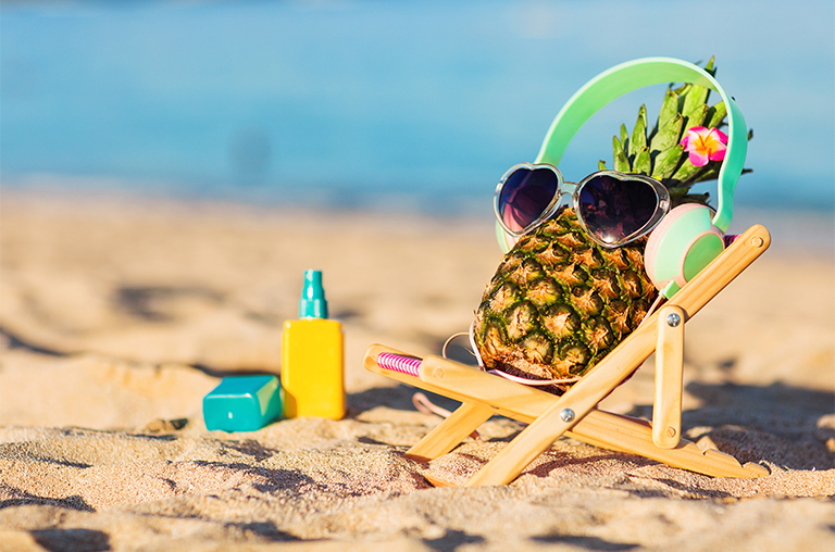 Eine Ananas drapiert mit einer Brille und Sonnenspray im Sand am Meer im strahlenden Sonnenschein